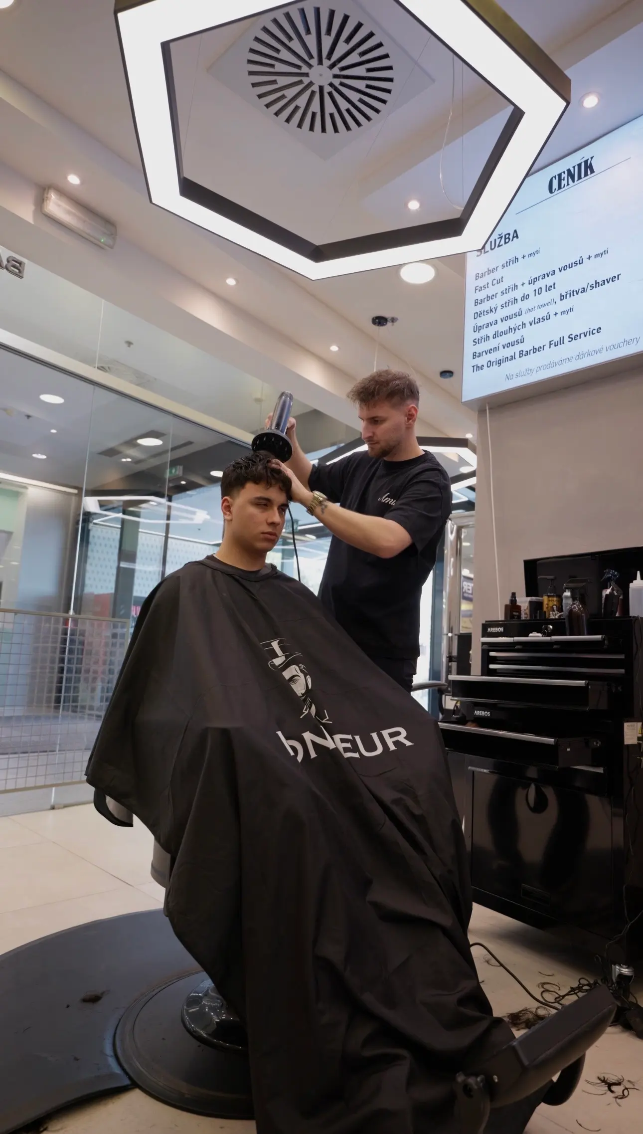 Barber fénuje klientovi vlasy vedle obrazovky s ceníkem služeb
