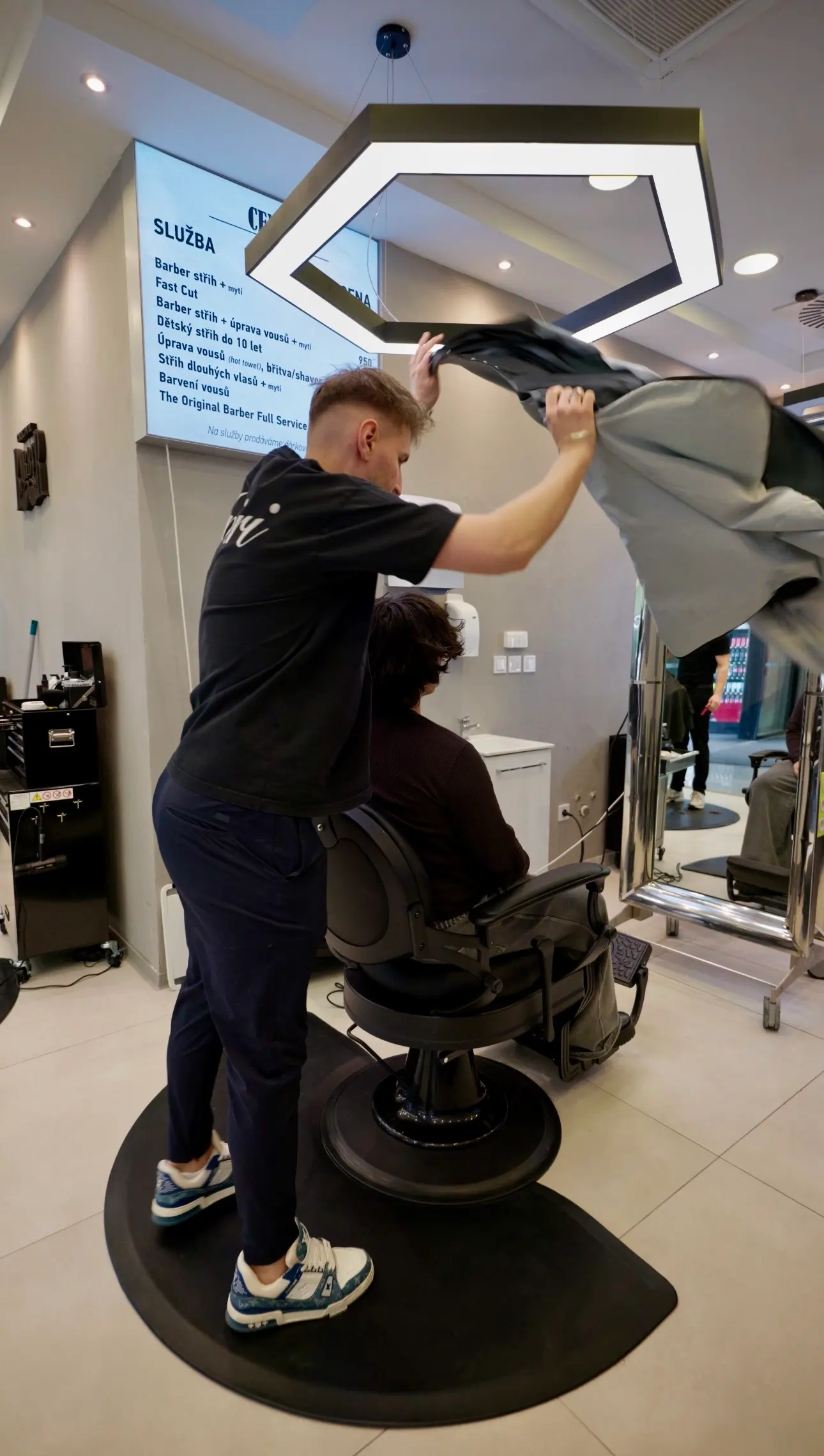Barber obléká klientovi pláštěnku před začátkem střihu